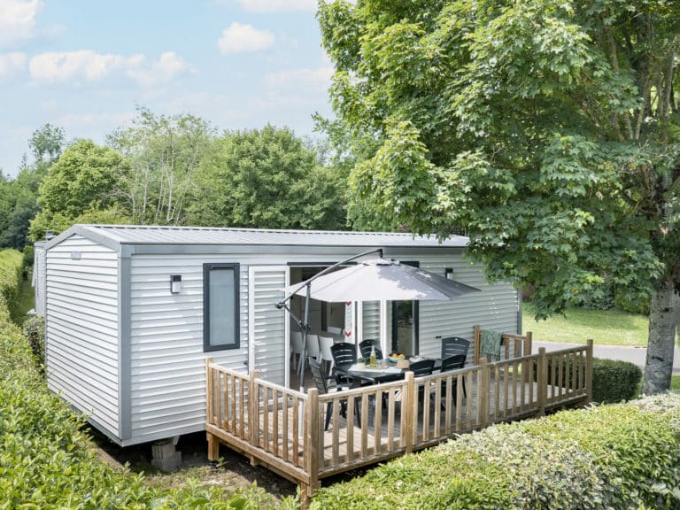 Mobil-home blanc avec terrasse et parasol, nature environnante.